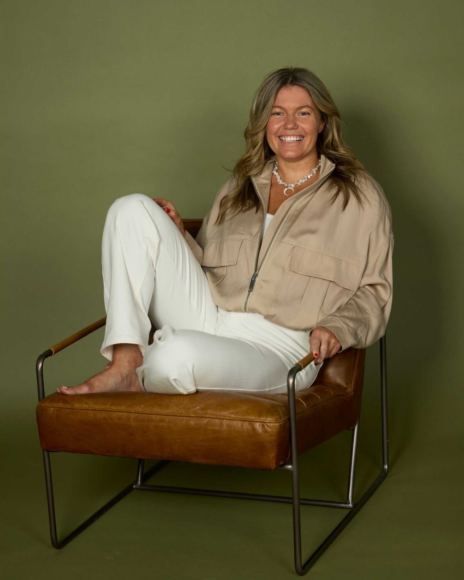 Olivia Carr sitting cross-legged in a leather armchair against an olive green background, wearing a beige jacket and white trousers, smiling at the camera.
