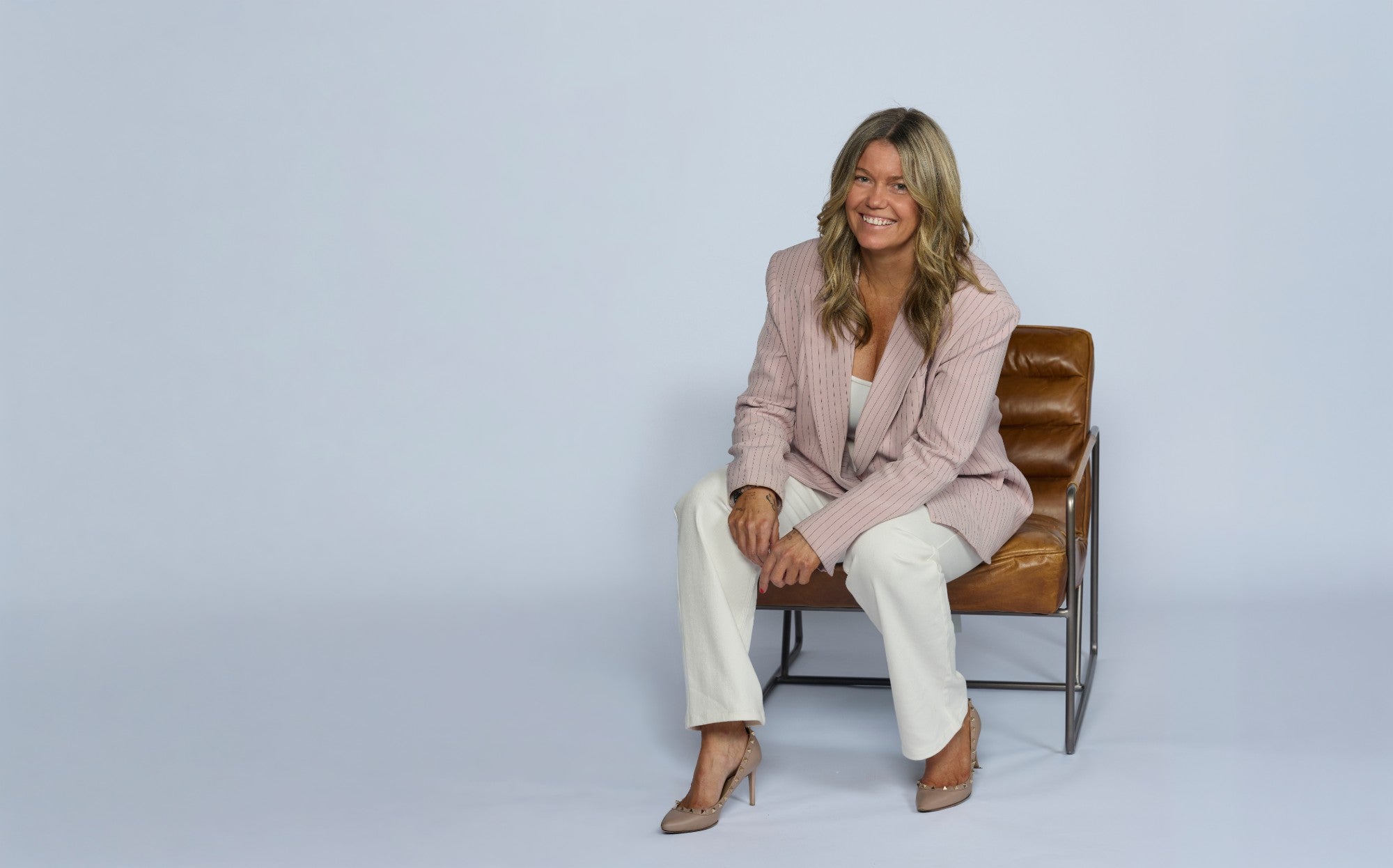 Olivia Carr seated in a pale pink pinstripe blazer and white trousers on a leather chair against a plain pale background, smiling at the camera.